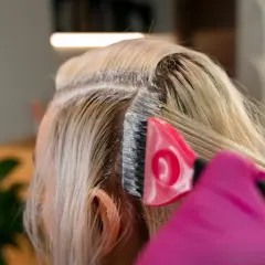 Profesjonalna dekoloryzacja włosów w salonie. Różowa rękawiczka i pędzel nakładają produkt na blond pasma.