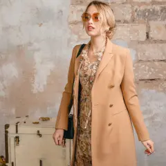Elegancka kobieta w płaszczu camelowy kolor, sukience w kwiaty i okularach przeciwsłonecznych pozuje przy starej walizce.