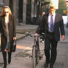 Kobieta w płaszczu i sukience, w stylu Carolyn Bessette, idzie z torebką. Obok niej mężczyzna w garniturze i czapce prowadzi rower.