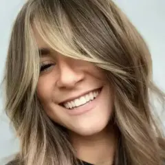 Uśmiechnięta kobieta z burzą **shaggy hair**, w blond i brązowych odcieniach, z grzywką zasłaniającą jedno oko.