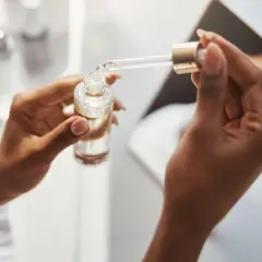 Dłonie trzymają buteleczkę z serum. Pipeta z kwasem ferulowym nabiera kroplę produktu.