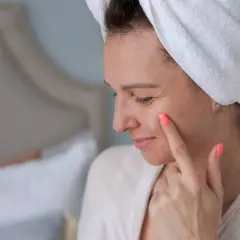 Kobieta z ręcznikiem na głowie aplikuje na twarz krem, być może z cennym olejem z czarnuszki.