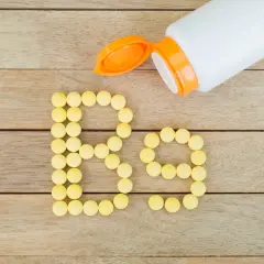 Żółte tabletki ułożone w napis "B9" obok otwartego białego pojemnika na drewnianym tle.