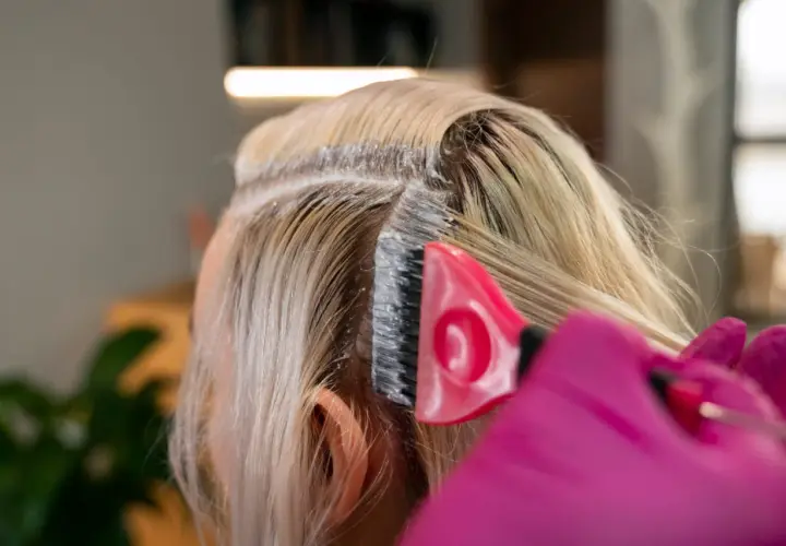 Profesjonalna dekoloryzacja włosów w salonie. Różowa rękawiczka i pędzel nakładają produkt na blond pasma.