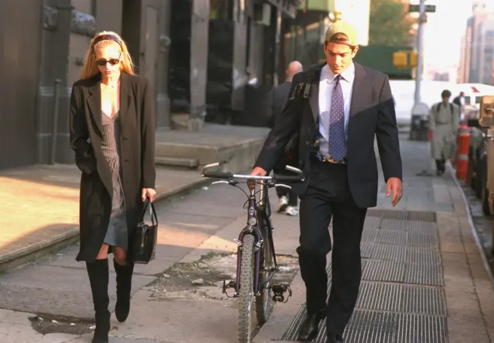 Kobieta w płaszczu i sukience, w stylu Carolyn Bessette, idzie z torebką. Obok niej mężczyzna w garniturze i czapce prowadzi rower.