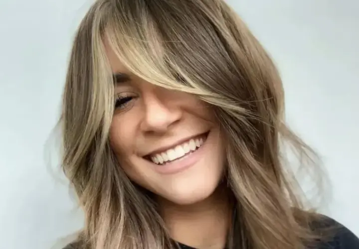 Uśmiechnięta kobieta z burzą **shaggy hair**, w blond i brązowych odcieniach, z grzywką zasłaniającą jedno oko.