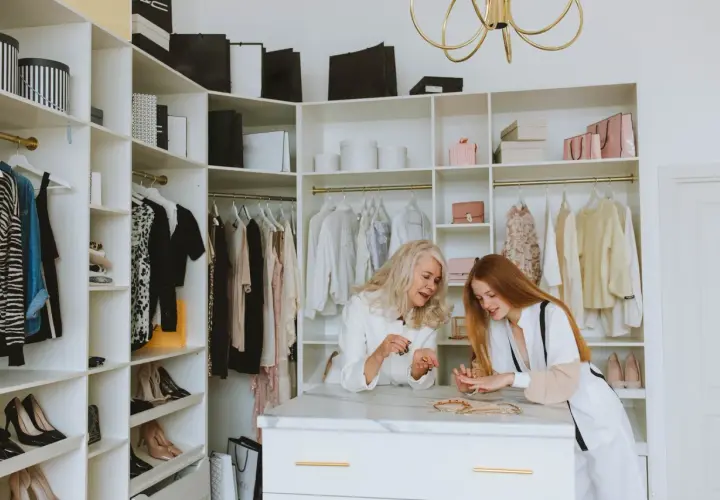 Dwie kobiety przeglądają biżuterię w garderobie. Ubrania i buty w stylu basic wiszą na wieszakach.