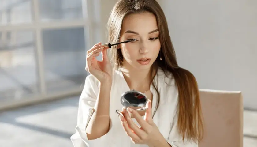 Młoda kobieta maluje rzęsy tuszem, który zawiera phenoxyethanol, patrząc w lusterko.