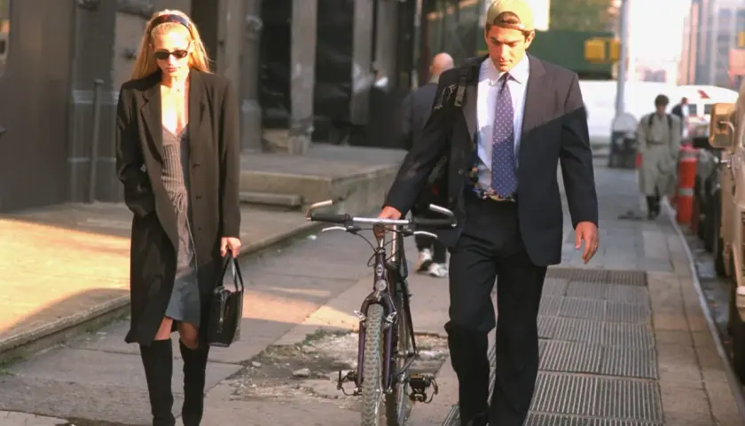 Kobieta w płaszczu i sukience, w stylu Carolyn Bessette, idzie z torebką. Obok niej mężczyzna w garniturze i czapce prowadzi rower.