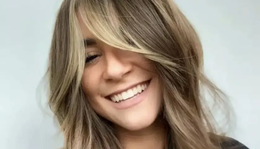 Uśmiechnięta kobieta z burzą **shaggy hair**, w blond i brązowych odcieniach, z grzywką zasłaniającą jedno oko.