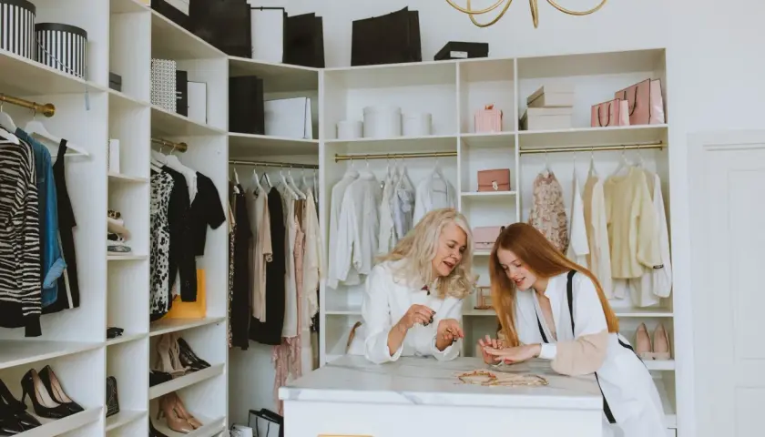 Dwie kobiety przeglądają biżuterię w garderobie. Ubrania i buty w stylu basic wiszą na wieszakach.