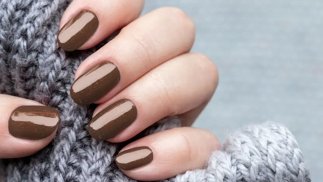 Dłonie z idealnym manicurem kombinowanym w odcieniu gorzkiej czekolady, otulone miękkim, szarym swetrem.