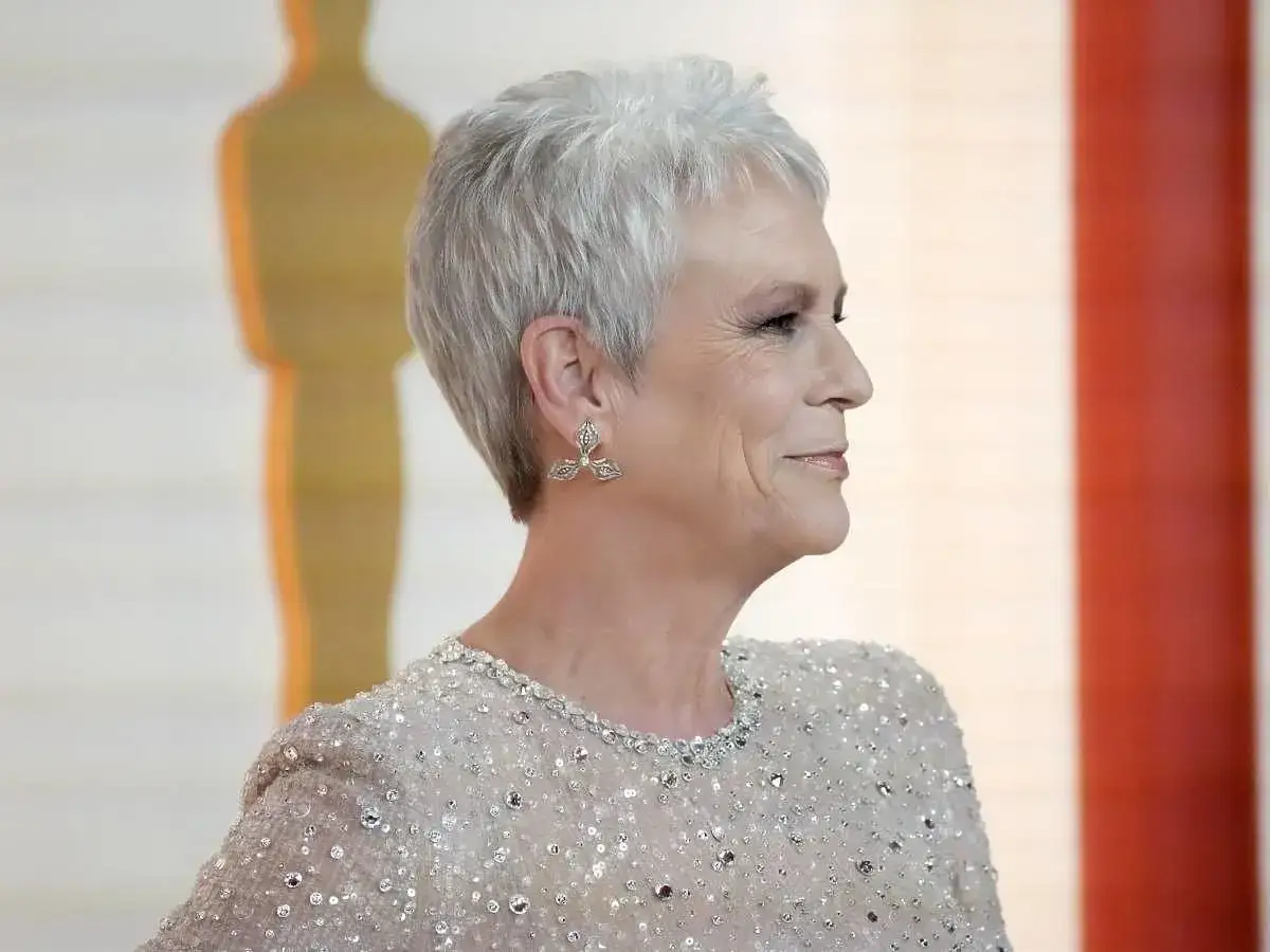 Elegancka, krótka fryzura dla pań po 50-tce, jak u Jamie Lee Curtis. Srebrne włosy, błyszcząca sukienka i stylowe kolczyki.