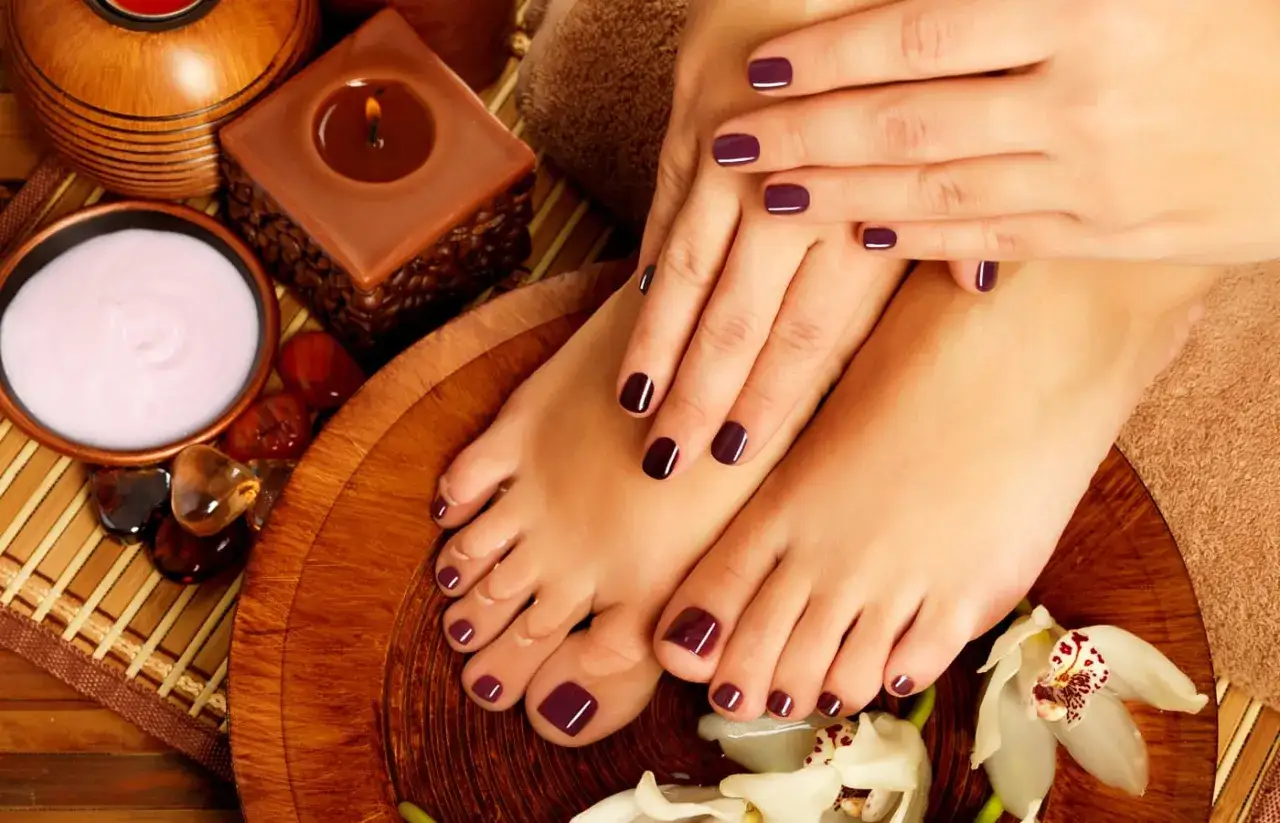 Relaksacyjny pedicure z ciemnofioletowymi paznokciami u nóg i dłoni. W tle świeca i krem do pielęgnacji.