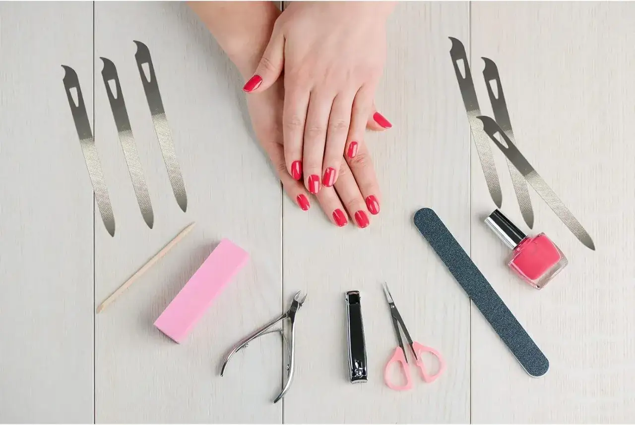 Dłonie z czerwonym lakierem, pilniki, cążki, nożyczki, polerka i różowy lakier – wszystko do wykonania idealnego manicure biologicznego.